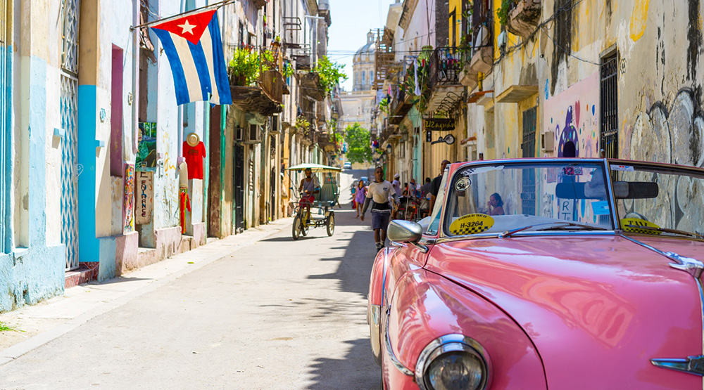 A street in Havana, Cuba. A pink car is in the foreground and the Cuban flag flies from a building, while people walk in the backround. The country has faced significant black market currency devaluation since Day Zero, when it reunified it currencies.